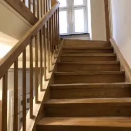 Jasne drewniane schody z balustradą, widok z dołu, naturalne oświetlenie z okna na górze.