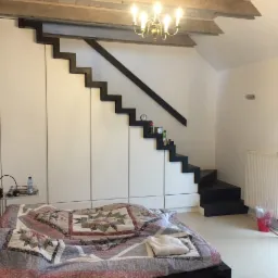 Biała szafa wnękowa z ukrytymi schodami prowadzącymi na górę, czarne schody tworzą kontrast z bielą szafy, w pomieszczeniu widoczna sypialnia z łóżkiem.