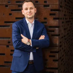 Mężczyzna w niebieskim garniturze, białej koszuli i brązowym pasku, stoi na tle rustykalnej, metalowej ściany z geometrycznymi wycięciami, prezentując profesjonalny wizerunek.