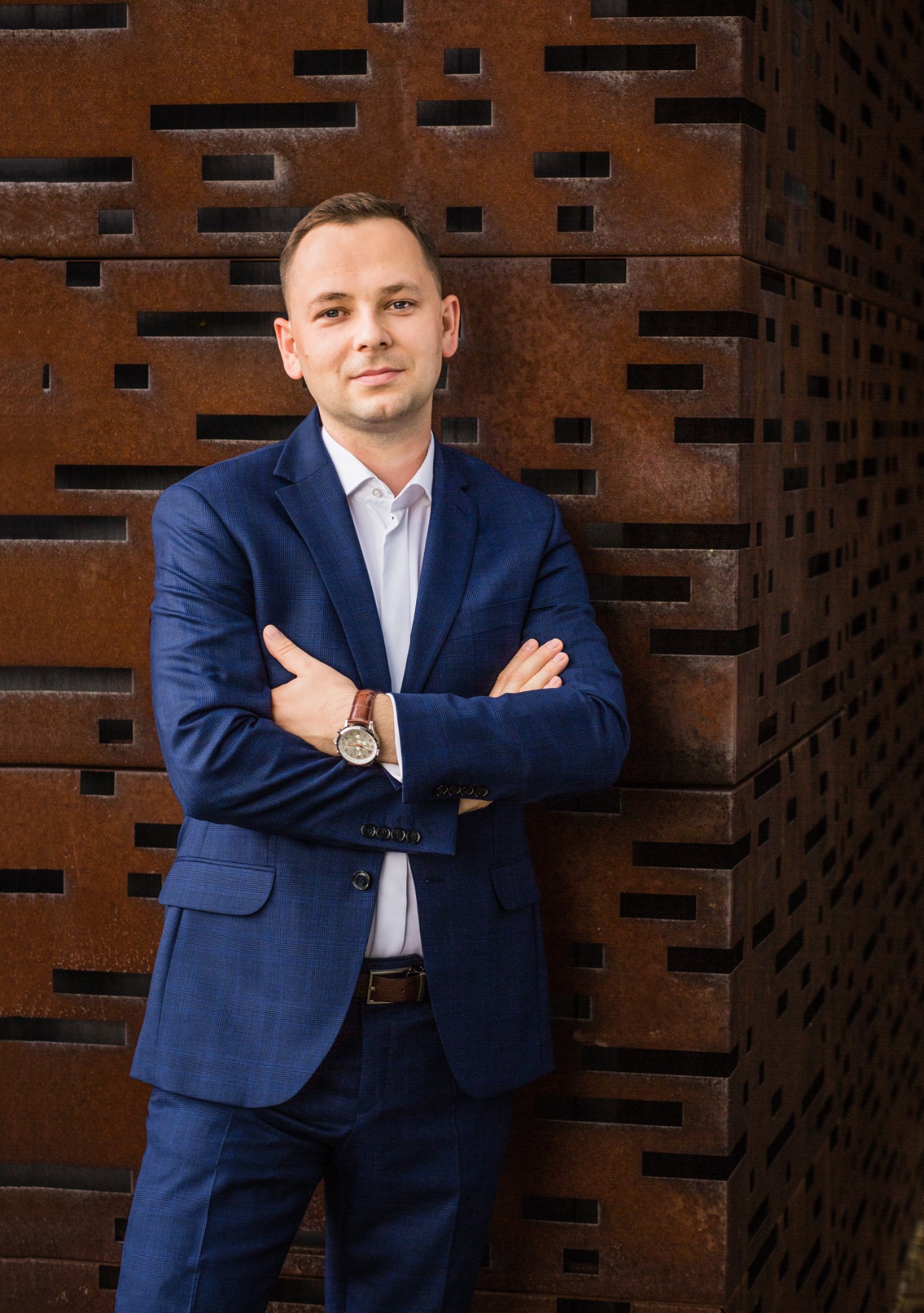 Mężczyzna w niebieskim garniturze, białej koszuli i brązowym pasku, stoi na tle rustykalnej, metalowej ściany z geometrycznymi wycięciami, prezentując profesjonalny wizerunek.