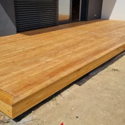 Nowoczesny taras z jasnego drewna, częściowo ukończony, z widoczną membraną i parą klapek pod spodem, w słoneczny dzień.