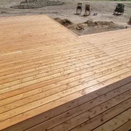 Nowo wybudowany drewniany taras przylegający do budynku, z widocznymi deskami ułożonymi w regularny wzór. W tle widoczne przygotowane podłoże, krzesła ogrodowe i narzędzia.