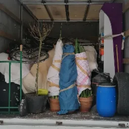 Wnętrze skrzyni ładunkowej wypełnione zabezpieczonymi przedmiotami podczas transportu, widoczne rośliny w doniczkach owinięte tkaniną i folią, rower, meble i inne przedmioty.