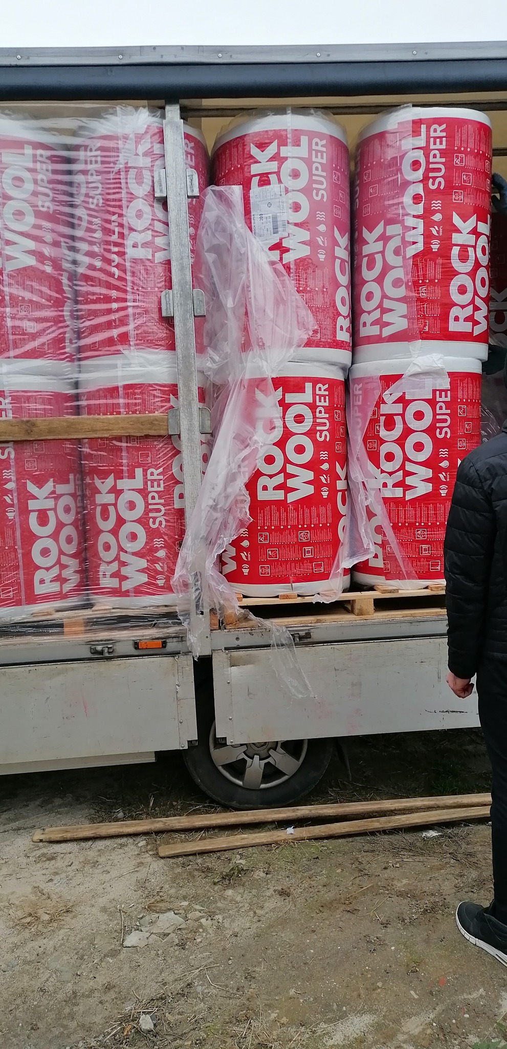 Załadunek rol izolacji Rockwool Super w opakowaniu foliowym na palecie do skrzyni ładunkowej samochodu dostawczego, widok z boku na koło i podwozie pojazdu.