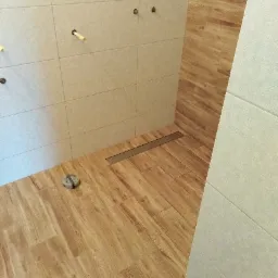 Wyłożona drewnopodobnymi płytkami podłoga i ściany w łazience z widocznym odpływem liniowym i niezamaskowanymi przyłączami hydraulicznymi.