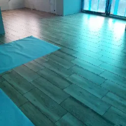 Ułożone panele podłogowe imitujące drewno, zabezpieczone fragmentami folii ochronnej, w surowym wnętrzu z oknem zasłoniętym folią malarską.