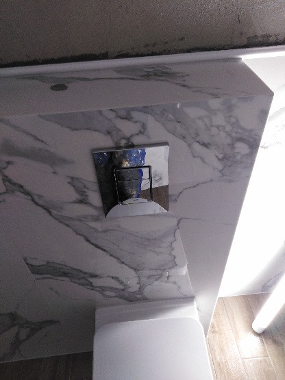 Szczegółowe ujęcie spłuczki WC zamontowanej na ścianie wyłożonej płytkami imitującymi marmur w odcieniach szarości, nad białą miską toaletową.