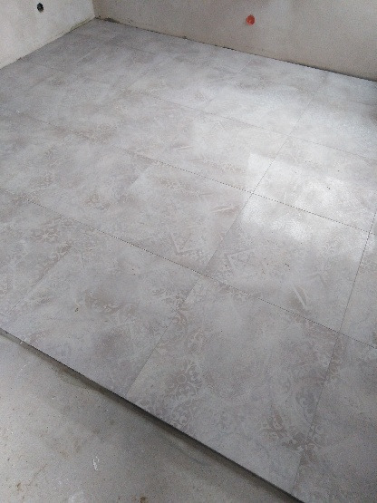 Świeżo ułożone, szare płytki podłogowe z delikatnym wzorem w pomieszczeniu w trakcie remontu, widoczny surowy beton na ścianach i wystające kable.