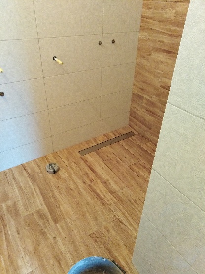 Wyłożona drewnopodobnymi płytkami podłoga i ściany w łazience z widocznym odpływem liniowym i niezamaskowanymi przyłączami hydraulicznymi.