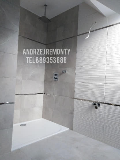 Nowoczesna łazienka z płytkami w odcieniach szarości i bieli, kabina prysznicowa typu walk-in z deszczownicą, minimalistyczna armatura, widoczne logo firmy na ścianie.