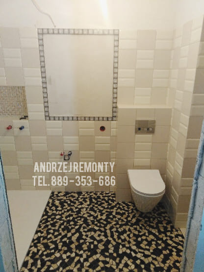 Wykończona łazienka z mozaikową podłogą w odcieniach czerni i beżu, kremowe płytki na ścianach, podwieszana toaleta i otwór rewizyjny obramowany dekoracyjnymi płytkami.