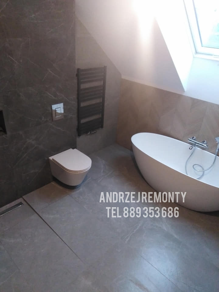 Nowoczesna łazienka na poddaszu z wolnostojącą wanną, wiszącą toaletą i grzejnikiem drabinkowym na ścianie wyłożonej szarym marmurem.