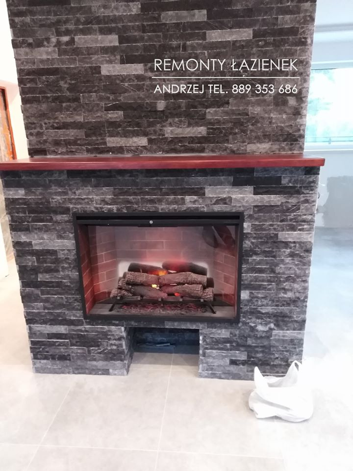 Obudowa kominka z szarej, łupanej cegły w łazience, z wkładem imitującym płomienie, zwieńczona ciemnobrązowym blatem, na podłodze jasne płytki, w tle widoczne okno i fragment glazury, w rogu leży...