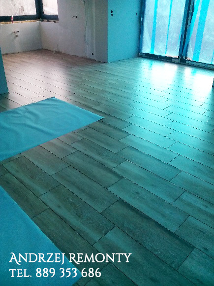 Ułożone panele podłogowe imitujące drewno, zabezpieczone fragmentami folii ochronnej, w surowym wnętrzu z oknem zasłoniętym folią malarską.