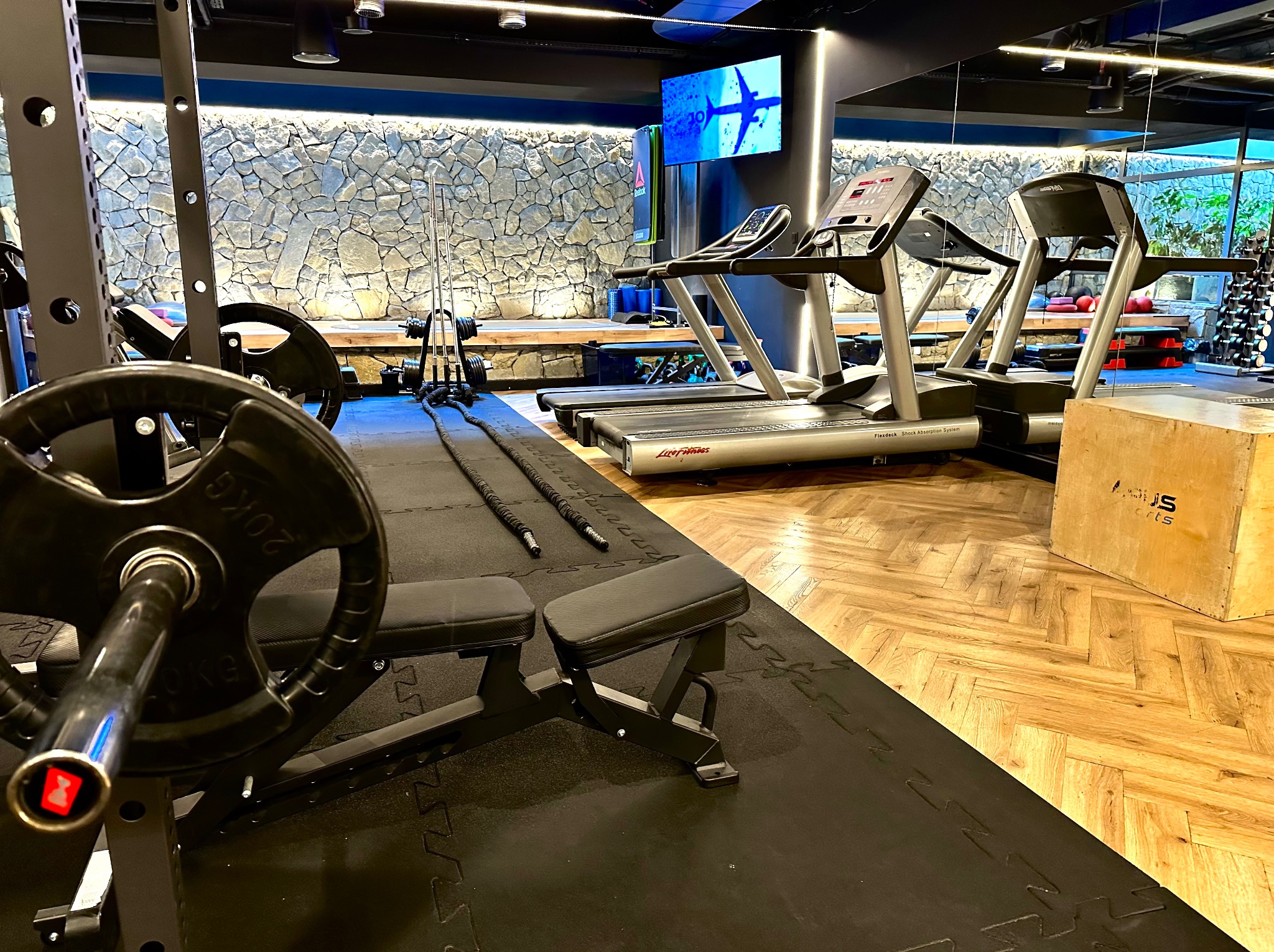 Nowoczesna siłownia z bieżniami, sztangą i ścianą z kamienia. Profesjonalny sprzęt do ćwiczeń, eleganckie wykończenie wnętrza, podłoga z gumy i drewna.