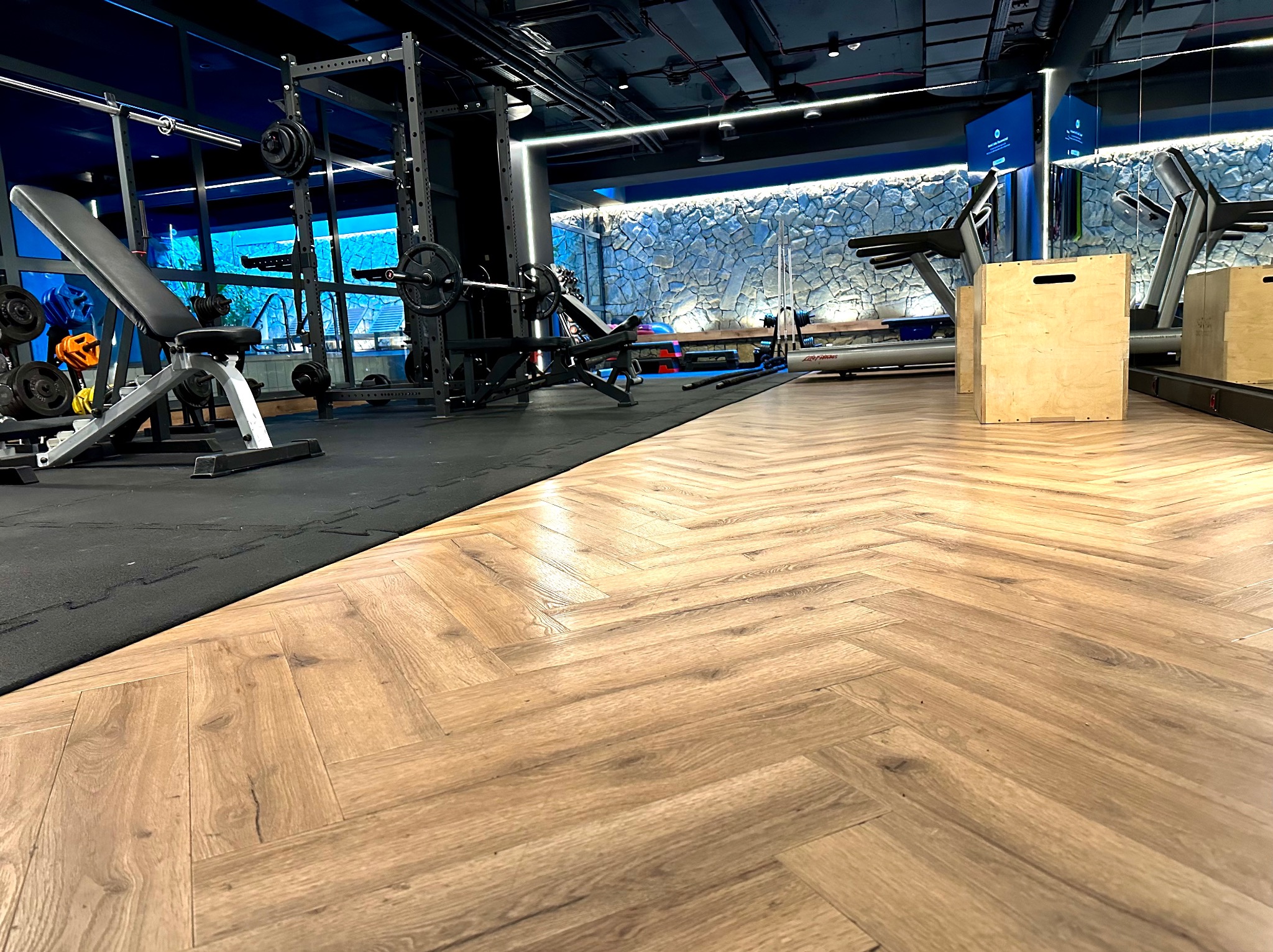 Nowoczesna siłownia z parkietem w jodełkę, sprzętem do ćwiczeń siłowych i bieżniami. Ściana wykończona kamieniem dekoracyjnym, oświetlenie LED.