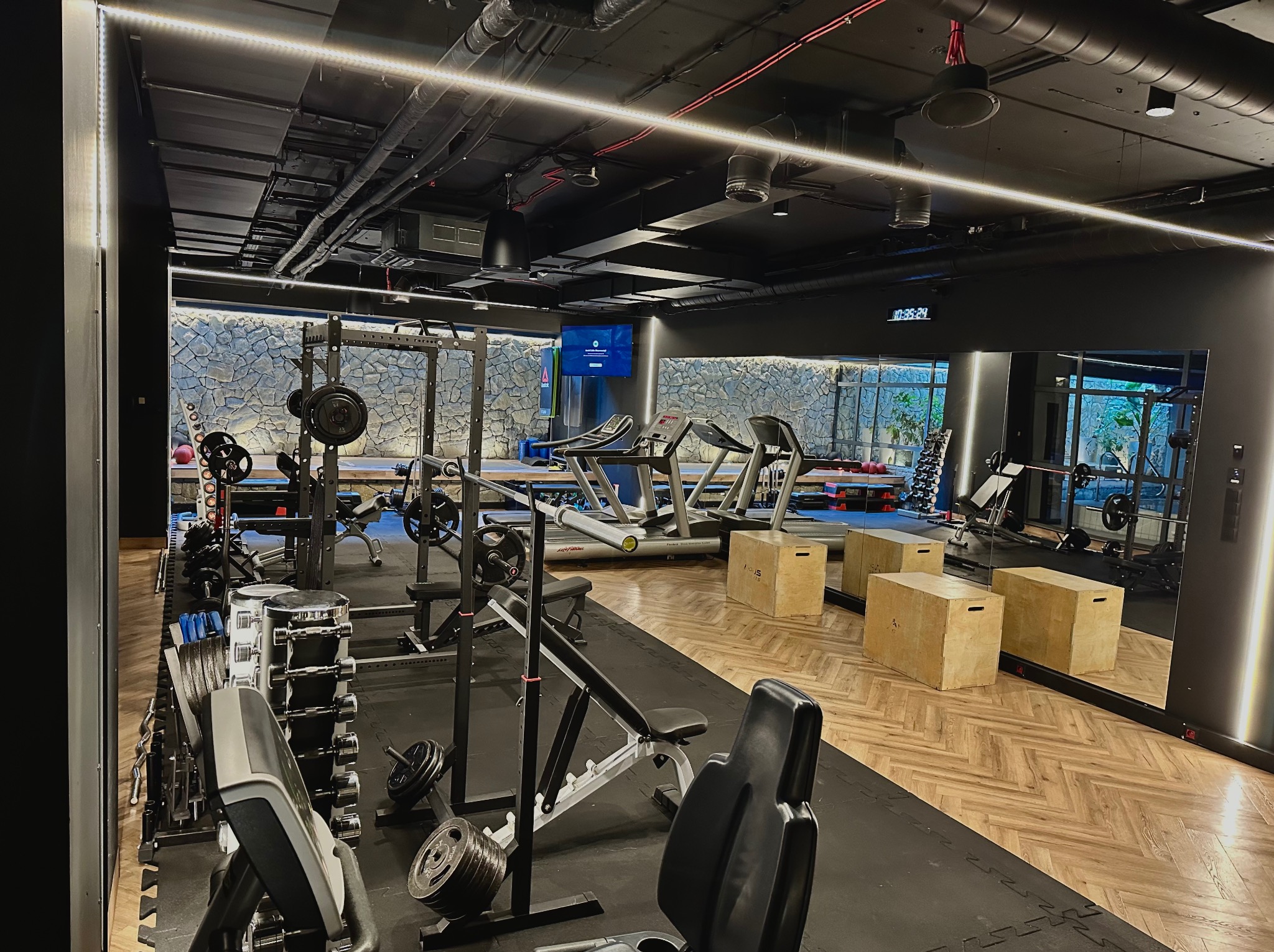 Profesjonalna siłownia z nowoczesnym sprzętem: bieżnie, ciężary, skrzynie do ćwiczeń. Ściana wykończona kamieniem, podłoga z parkietu. Duże lustra optycznie powiększają przestrzeń.