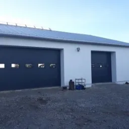 Dwie ciemnoszare segmentowe bramy garażowe z oknami w białym budynku z szarą blachą falistą na dachu, widok z zewnątrz na żwirowym podłożu.