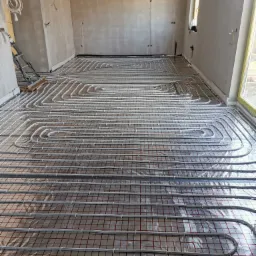 Białe rury ogrzewania podłogowego ułożone na srebrnej folii z czerwoną kratką w surowym wnętrzu z oknem i drabiną.