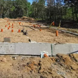 Fundament domu w trakcie budowy z wystającymi rurami kanalizacyjnymi, widok z bliska na betonową konstrukcję i przygotowany teren pod wylewkę, w tle las.