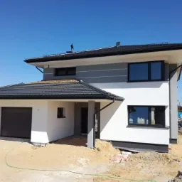 Nowoczesny dom jednorodzinny z szarą elewacją, czarną stolarką okienną i bramą garażową, w trakcie prac wykończeniowych terenu.