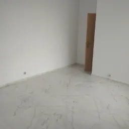 Puste, odnowione pomieszczenie z białymi ścianami i podłogą wyłożoną jasnymi, marmurkowymi płytkami. Widoczny fragment drewnianych drzwi z klamką i gniazdka elektryczne na ścianach.