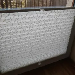 Zamknięta, biała, plisowana osłona okienna z delikatnym, szarym wzorem liści na balkonie.