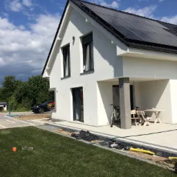 Nowoczesny dom jednorodzinny z białą elewacją, czarną stolarką okienną i panelami słonecznymi na dachu, widok z ogrodu w słoneczny dzień, z widocznymi pracami wykończeniowymi wokół budynku.