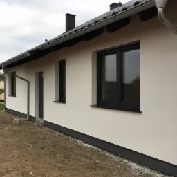 Elewacja budynku jednorodzinnego po tynkowaniu, z widocznymi oknami w ciemnych ramach, orynnowaniem i czarnym pasem izolacji przy ziemi.