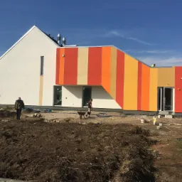 Nowoczesny dom z pomalowaną elewacją w pionowe pasy w odcieniach pomarańczy, żółci i beżu, widoczny pracownik z taczką oraz drugi pracownik oddalający się od budynku, teren wokół w trakcie prac...