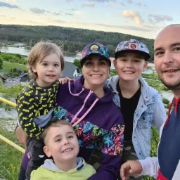 Rodzinne zdjęcie z trzema synami na tle malowniczego krajobrazu z jeziorem i lasem, zrobione z lekkiego wzniesienia przy żółtej metalowej barierce.