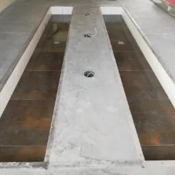 Wnętrze w trakcie wykańczania z dwoma wgłębieniami wyłożonymi ciemnymi płytkami, oddzielonymi szarą, betonową platformą z otworami na instalacje; widoczne przewody, płytki na podłodze i fragment...