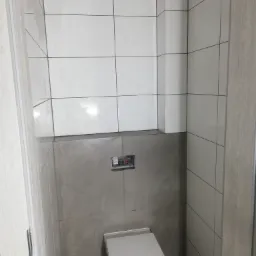 Nowoczesna toaleta podwieszana z białą ceramiką i szarą obudową, widoczna częściowo zabudowana spłuczka, białe płytki na ścianach powyżej obudowy, szare płytki poniżej, drzwi z okleiną imitującą...