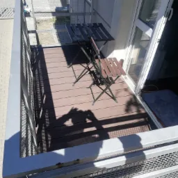 Umeblowany balkon z brązową podłogą z desek kompozytowych, metalową balustradą i składanym stolikiem z krzesłem.