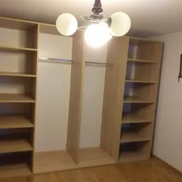 Jasna, drewniana szafa wnękowa z otwartymi półkami i drążkami na ubrania, oświetlona trzypunktową lampą sufitową, na tle białej ściany z obrazkiem.