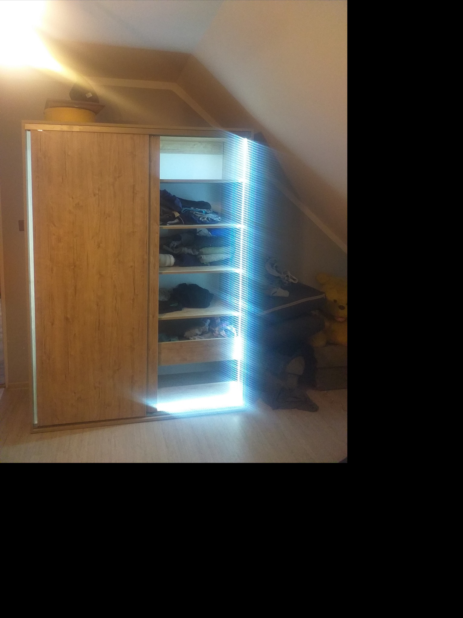 Drewniana szafa przesuwna z oświetleniem LED wewnątrz, częściowo otwarta, ukazująca półki z ubraniami w pokoju na poddaszu.