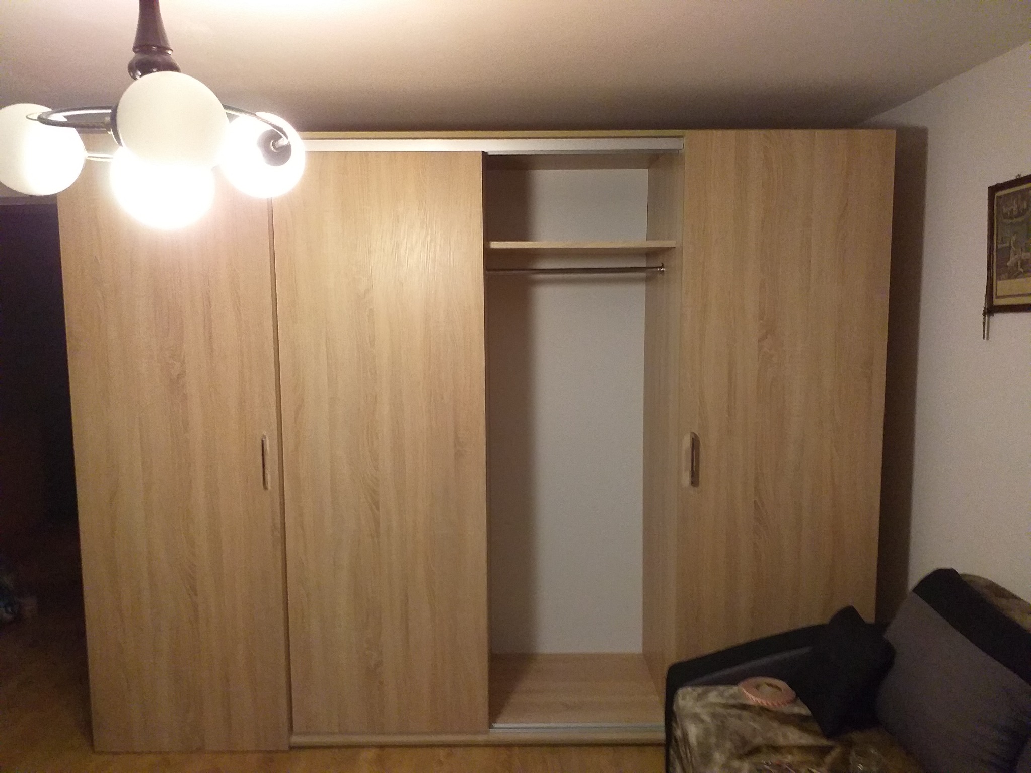 Jasna szafa przesuwna z drewnopodobnymi frontami, częściowo otwarta, z widoczną półką i drążkiem, oświetlona przez lampę sufitową z kulistymi kloszami.