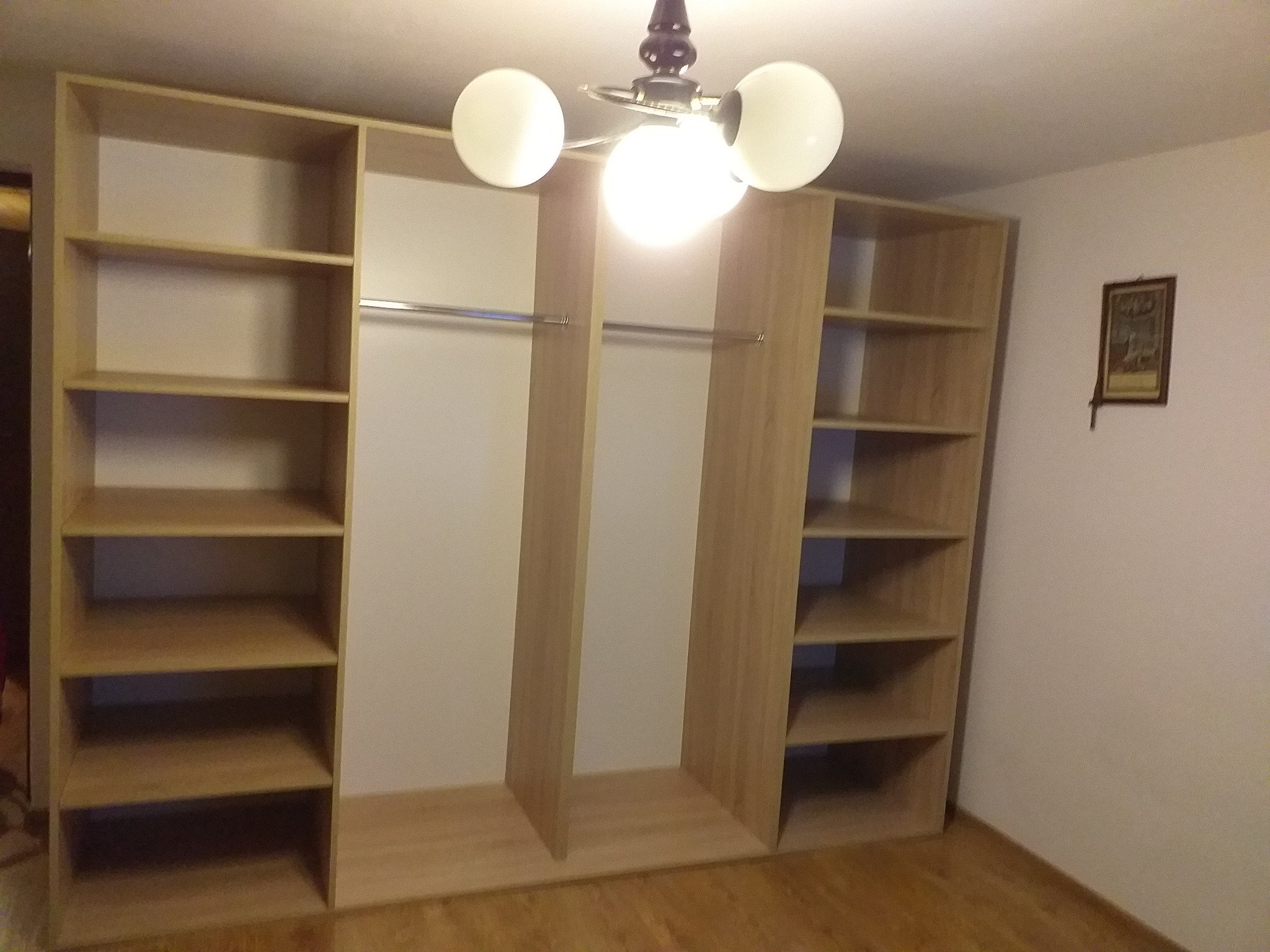 Jasna, drewniana szafa wnękowa z otwartymi półkami i drążkami na ubrania, oświetlona trzypunktową lampą sufitową, na tle białej ściany z obrazkiem.