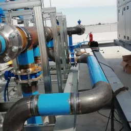 Instalacja przemysłowa na dachu budynku: rurociąg z rur stalowych, częściowo pomalowanych na niebiesko, z widocznymi spawami, połączony za pomocą kołnierzy, wsparty na metalowej konstrukcji. W tle...