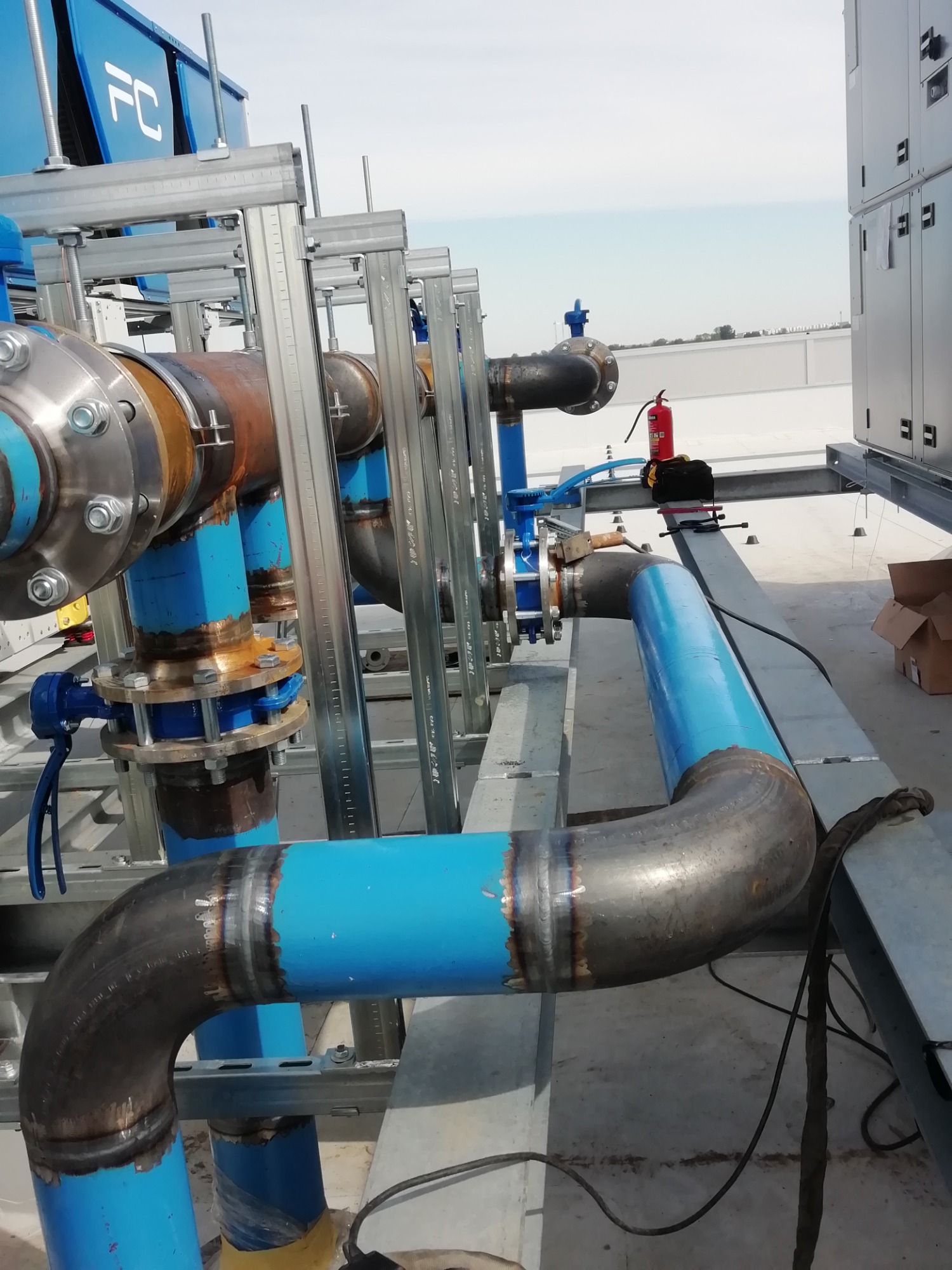 Instalacja przemysłowa na dachu budynku: rurociąg z rur stalowych, częściowo pomalowanych na niebiesko, z widocznymi spawami, połączony za pomocą kołnierzy, wsparty na metalowej konstrukcji. W tle...