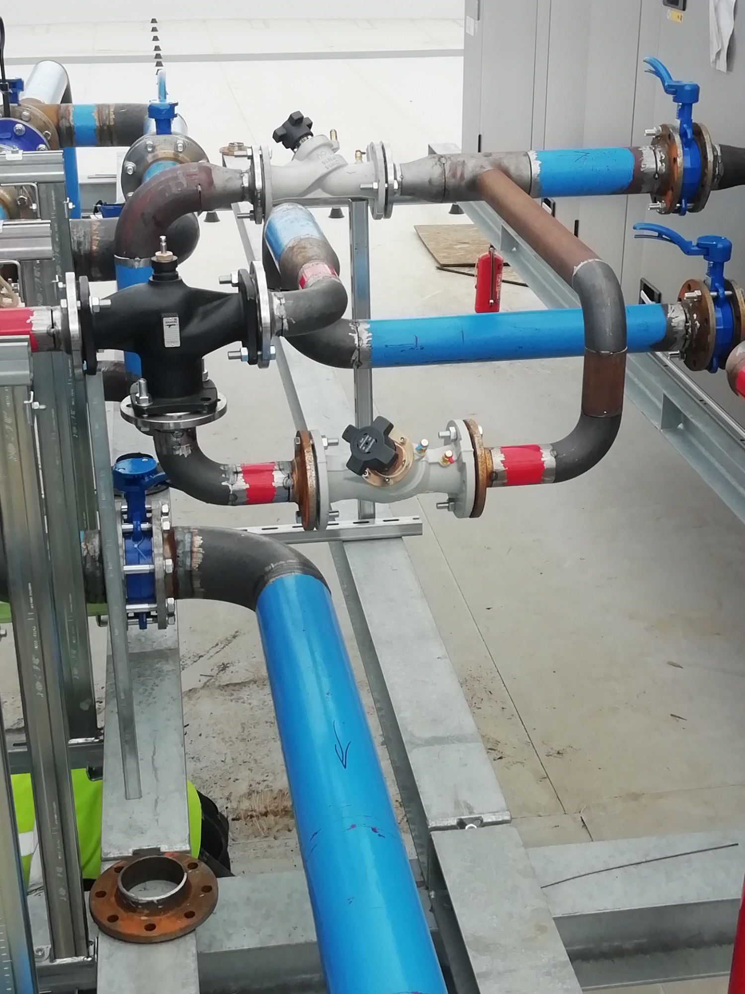 Złożona instalacja rur z zaworami i flanszami, malowana w kolorach niebieskim i czerwonym, częściowo spawana, zamontowana na metalowej konstrukcji wsporczej.