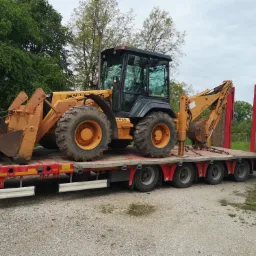 Żółto-czarna koparko-ładowarka CASE 595 na czerwonej niskopodwoziowej platformie transportowej, zabezpieczona łańcuchami, z mężczyzną w szarej bluzie przy przedniej łyżce, na tle zieleni...