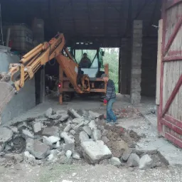 Żółta koparko-ładowarka CASE z łyżką nabierającą gruz na tle otwartej stodoły z dwoma pracownikami, jeden w kabinie maszyny, drugi w czapce i czerwonych rękawicach stojący obok sterty gruzu.