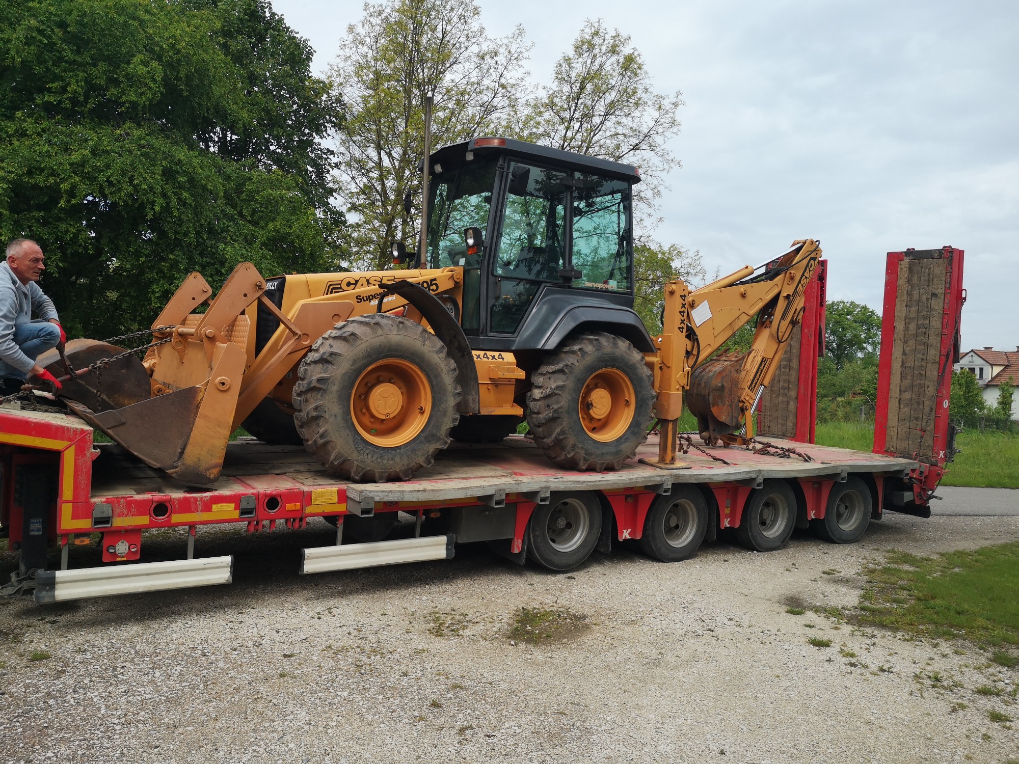Żółto-czarna koparko-ładowarka CASE 595 na czerwonej niskopodwoziowej platformie transportowej, zabezpieczona łańcuchami, z mężczyzną w szarej bluzie przy przedniej łyżce, na tle zieleni...