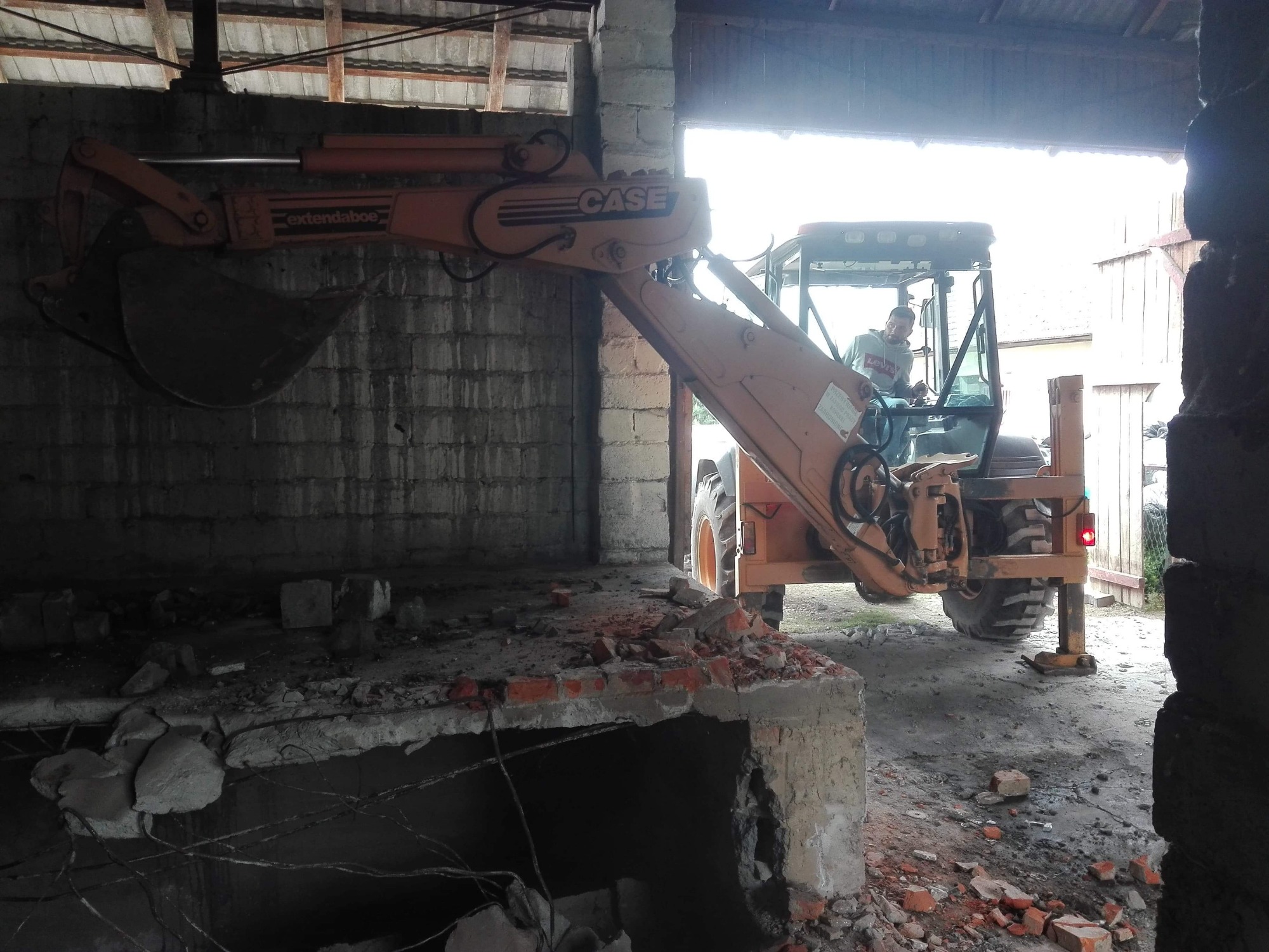 Pomarańczowa koparko-ładowarka CASE z wysuniętym ramieniem i łyżką, częściowo wewnątrz budynku, gruz ceglany na pierwszym planie, operator w kabinie, widoczne elementy konstrukcyjne budynku.
