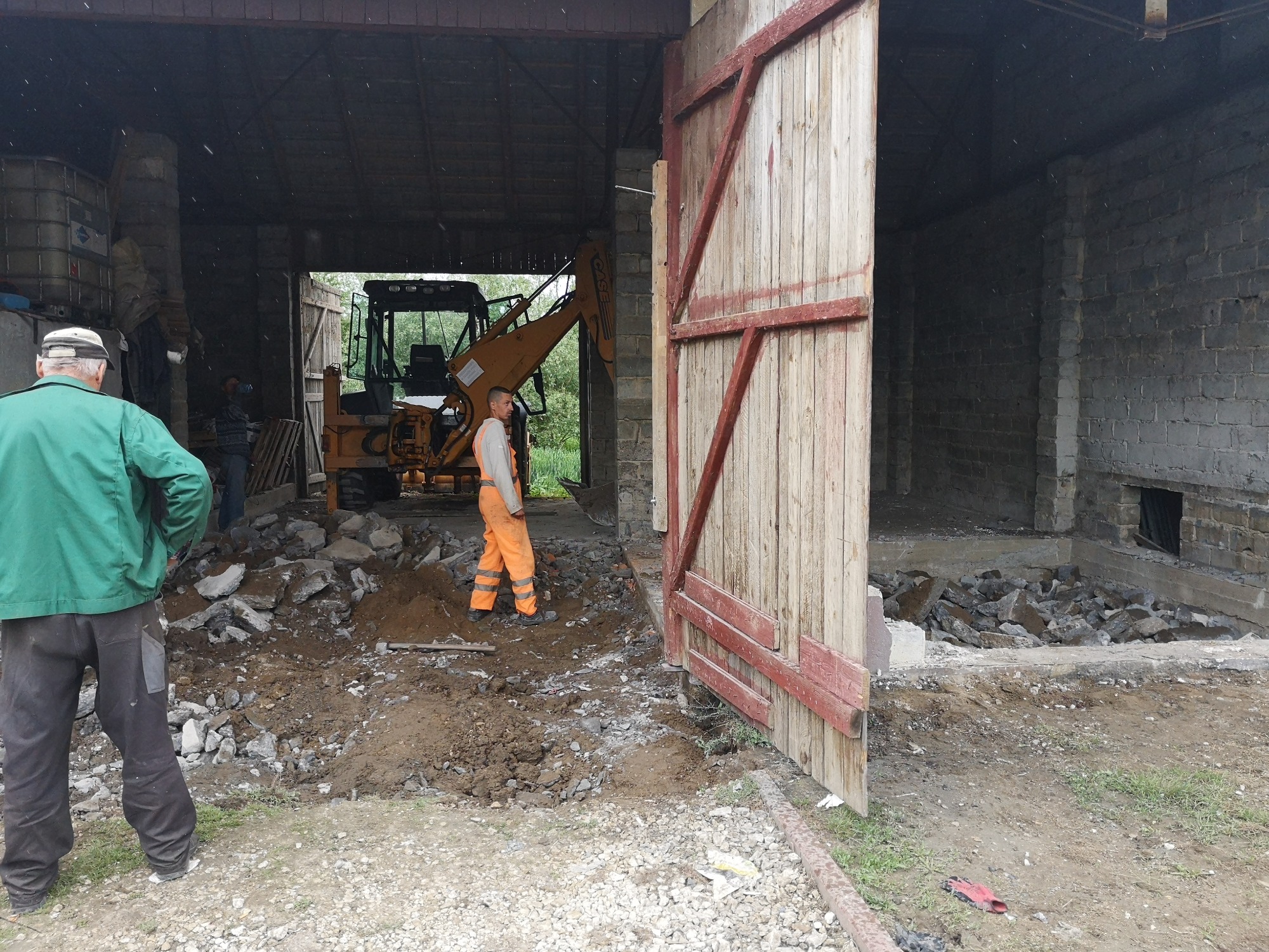 Robotnik w pomarańczowym kombinezonie stoi przed otwartymi drewnianymi wrotami, w tle koparka i gruzowisko po wyburzeniu fragmentu ściany budynku z szarej cegły.