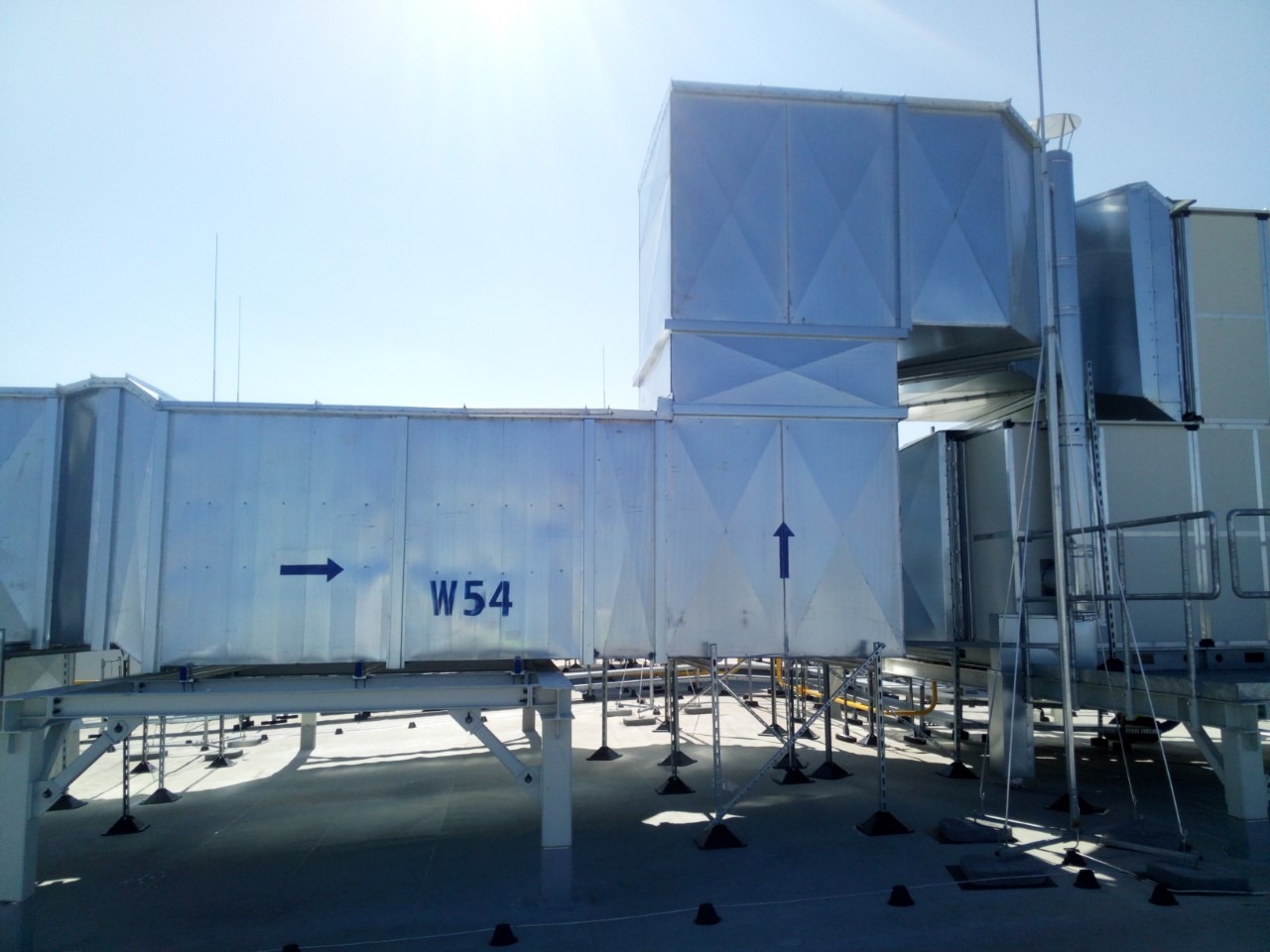 System wentylacyjny z metalowych kanałów na dachu budynku komercyjnego, oznaczony strzałkami kierunku przepływu powietrza i numerem W54, wsparty na konstrukcji stalowej z regulowanymi stopkami.