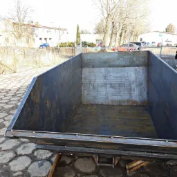 Nowy, spawany kontener do wywozu odpadów, umieszczony na drewnianych belkach, z widocznymi spawami i surową powierzchnią metalu na tle betonowego placu i zabudowań.