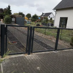 Dwuskrzydłowa, grafitowa brama panelowa z widoczną skrzynką na listy z numerem 3, prowadząca na podjazd z kamiennym wypełnieniem, w tle dom z ogrodem i placem zabaw.