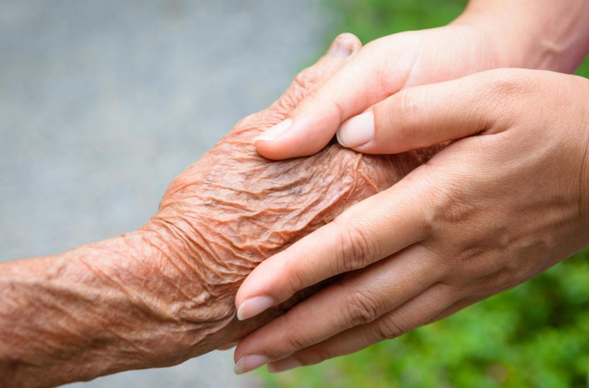 Dłoń osoby starszej spoczywająca w dłoni osoby młodszej, ukazująca troskę i wsparcie.